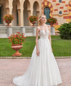 Denna glamorösa balinspirerade brudklänning kombinerar romantisk design med en touch av drama. Klänningen är sydd i glittrande tyll och har en axelbandslös, transparent livstycke med en djup hjärtformad urringning och eleganta av axeln-band. Det strukturerade livstycket pryds av synlig korsettliknande boning som ger en skulpterande effekt, medan paljettbroderade spetsapplikationer i bomull sveper från bodice ner över kjolen i en mjuk, nedtonad övergång. Glittertyllklädda knappar löper längs ryggen och en lätt svepande släp (sweep train) ger klänningen en graciös avslutning. En perfekt kombination av sagolik romantik och modern glamour.